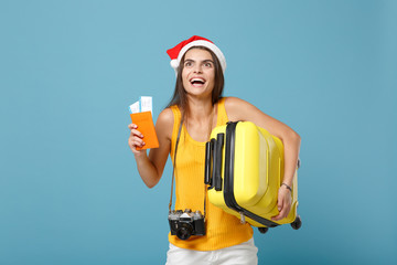 Traveler tourist woman in Santa hat, yellow casual clothes hold tickets bag camera isolated on blue background. Female passenger traveling abroad travel on weekends getaway. Air flight journey concept