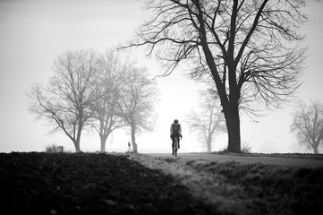 Black and white photo of a man gravel bike. Cyclist on the empty road and the trees in the mist.