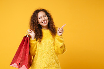 Smiling african american girl posing isolated on yellow orange background. People lifestyle concept. Mock up copy space. Holding package bag with purchases after shopping, pointing index finger aside.