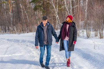 Handsome man and attractive young woman walking along snowy country road in sunny day. Beautiful look, male and female fashion, winter outfit. Winter holidays, weekend at countryside concept