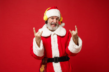 Fototapeta na wymiar Elderly gray-haired mustache bearded Santa man in Christmas hat posing isolated on red background. New Year 2020 celebration concept. Mock up copy space. Listen music with headphones showing horns up.