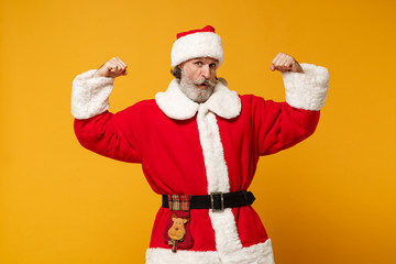 Fototapeta na wymiar Strong elderly gray-haired mustache bearded Santa man in Christmas hat posing isolated on yellow wall background. Happy New Year 2020 celebration concept. Mock up copy space. Showing biceps, muscles.