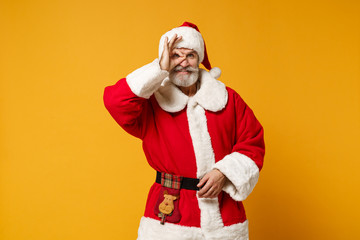 Fototapeta na wymiar Elderly gray-haired mustache bearded Santa man in Christmas hat isolated on yellow background. New Year 2020 celebration concept. Mock up copy space Hold hand near eye imitating binoculars or glasses.