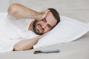 Fototapeta na wymiar Sad young handsome bearded man lying in bed with white sheet pillow blanket in bedroom at home. Tired upset male spending time in room. Rest relax good mood lifestyle concept. Mock up copy space.