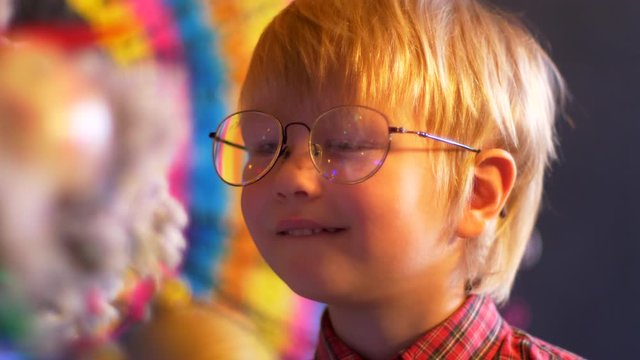 Cute blond boy in plaid shirt and tie in big eyeglasses sits next to Christmas tree on colored background. glasses reflect flashing garlands with bright lights. Shallow focus with old movie effect