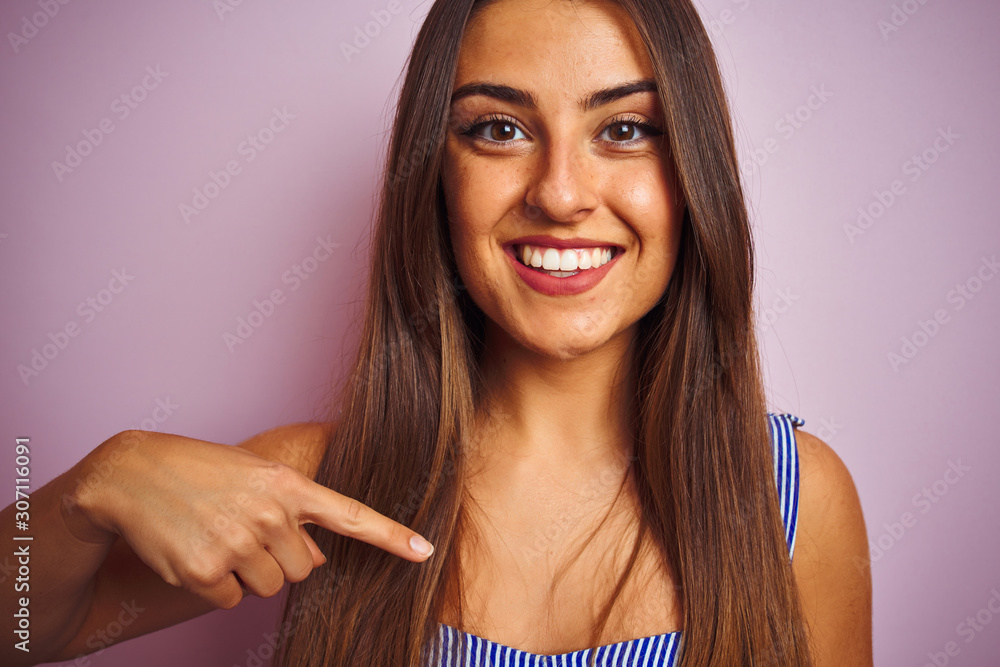 Wall mural Young beautiful woman wearing t-shirt standing over isolated pink background with surprise face pointing finger to himself