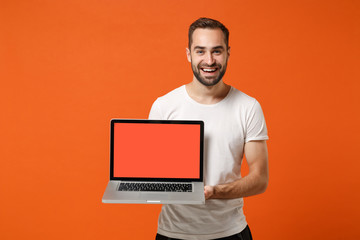 Smiling young man in casual white t-shirt posing isolated on bright orange background studio portrait. People lifestyle concept. Mock up copy space. Holding laptop pc computer with blank empty screen.