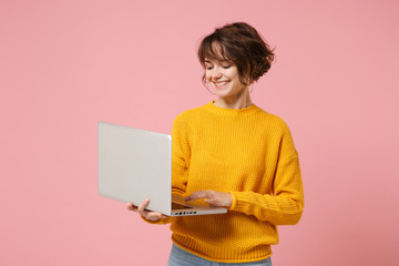 Smiling young brunette woman girl in yellow sweater posing isolated on pastel pink background studio portrait. People lifestyle concept. Mock up copy space. Holding and working on laptop pc computer. - obrazy, fototapety, plakaty
