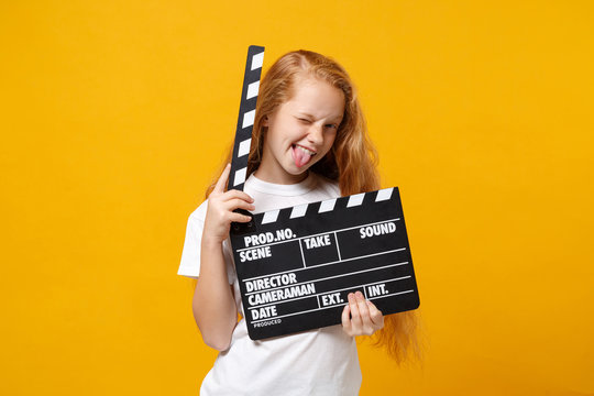 Little ginger kid girl 12-13 years old in white t-shirt isolated on yellow background. Childhood lifestyle concept. Mock up copy space. Holding classic black film making clapperboard, showing tongue.