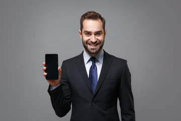 Smiling young business man in classic suit shirt tie posing isolated on grey background. Achievement career wealth business concept. Mock up copy space. Holding mobile phone with blank empty screen.