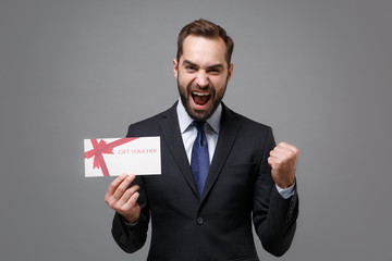 Joyful young business man in classic suit shirt tie posing isolated on grey background. Achievement career wealth business concept. Mock up copy space. Holding gift certificate, doing winner gesture.