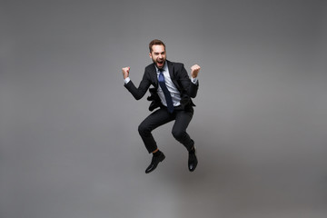 Joyful young business man in classic black suit shirt tie posing isolated on grey background. Achievement career wealth business concept. Mock up copy space. Jumping, doing winner gesture, screaming.