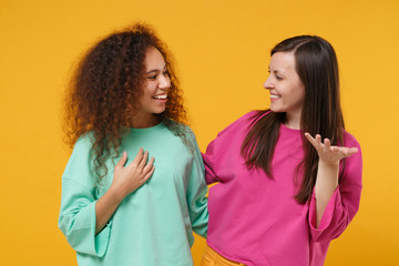 Two smiling cheerful women friends european and african american girls in pink green clothes posing isolated on yellow background. People lifestyle concept. Mock up copy space. Looking at each other.