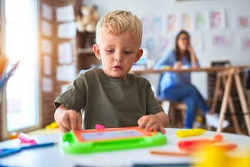 Young caucasian child playing at playschool with teacher. Mother and son at playroom drawing a draw with color pencils, young woman at the background sitting on desk.