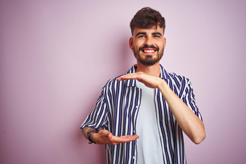 Young man with tattoo wearing striped shirt standing over isolated pink background gesturing with hands showing big and large size sign, measure symbol. Smiling looking at the camera. Measuring 