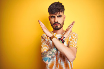 Young man with tattoo wearing striped t-shirt standing over isolated yellow background Rejection expression crossing arms doing negative sign, angry face