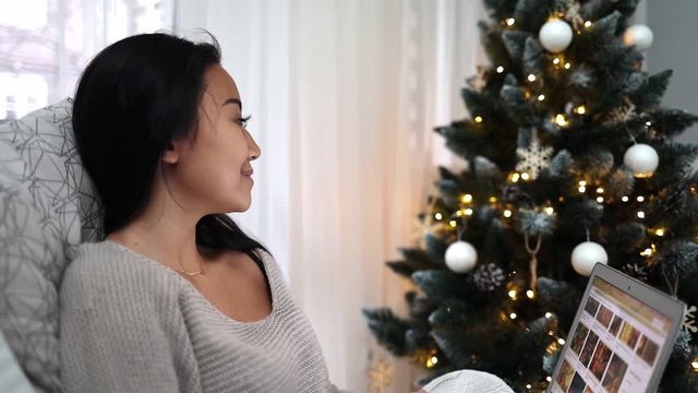 Happy asian girl shopping online with laptop sitting at decorated christmas tree background