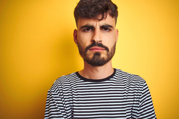 Obraz premium Young man with tattoo wearing striped t-shirt standing over isolated yellow background skeptic and nervous, disapproving expression on face with crossed arms. Negative person.