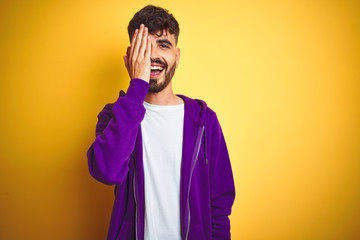 Young man with tattoo wearing sport purple sweatshirt over isolated yellow background covering one eye with hand, confident smile on face and surprise emotion.