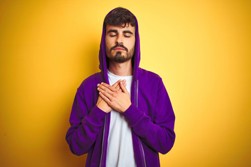 Young man with tattoo wearing purple sweatshirt with hood over isolated yellow background smiling with hands on chest with closed eyes and grateful gesture on face. Health concept.