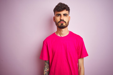 Young man with tattoo wearing t-shirt standing over isolated pink background with serious expression on face. Simple and natural looking at the camera.