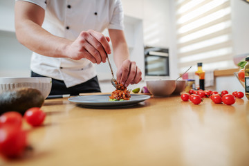 Cropped picture of dedicated caucasian chef standing in kitchen and putting cooked salmon on plate. On kitchen counter are different sorts of vegetables.