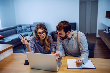 Young cheerful couple sitting at dining table and using laptop for online shopping. Man pointing at monitor and writing down things that he want while woman holding credit card.