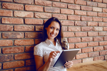 Fototapeta premium Stunning brunette in shirt sitting in cafe, leaning on brick wall and using tablet while looking at camera.