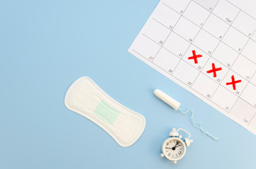 Flat lay female cycle calendar with menstrual pad, tampon and white alarm clock on a pastel background