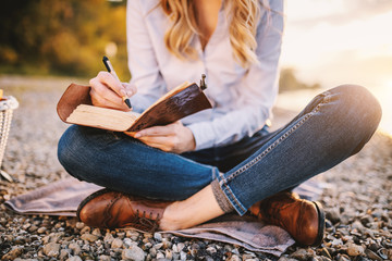 Cropped picture of focused fashionable beautiful caucasian woman sitting near river and holding notebook. - obrazy, fototapety, plakaty