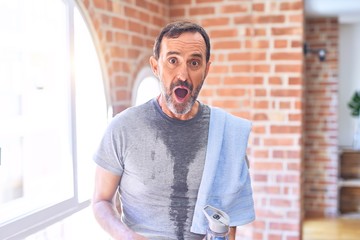 Middle age handsome sportman sweaty holding bottle of water and towel after exercise at gym scared...