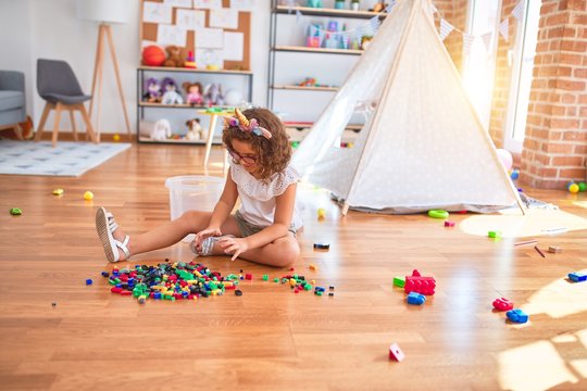 Beautiful toddler wearing glasses and unicorn diadem sitting on the floor playing with building blocks at kindergarten