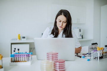 Beautiful caucasian female laboratory assistant sitting in lab and typing on laptop blood test...