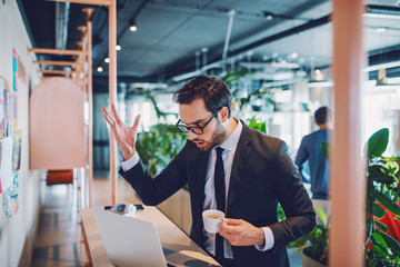 Frustrated handsome caucasian bearded businessman in suit and with eyeglasses holding cup with coffee and looking at laptop. Company interior.