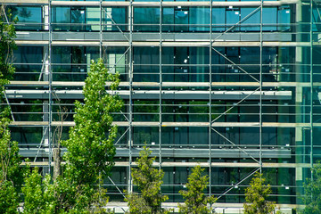 Construction site of modern glass building near the city park. Glass facade, scaffolds, and green...