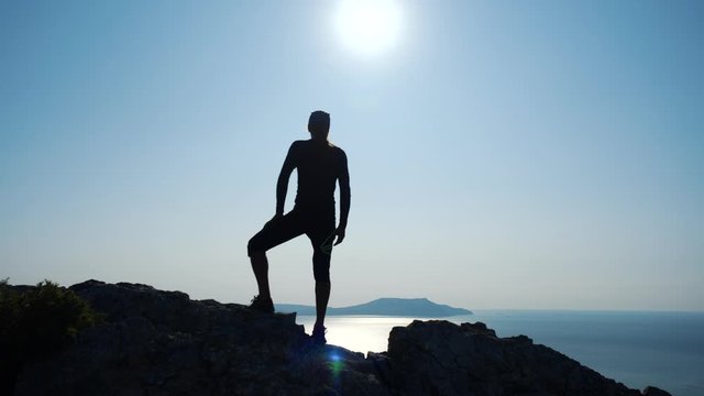 Young inspired man traveller is standing on the top of a mountain above the sea against beautiful blue sky. Silhouette of a happy hiker man standing on the summit.