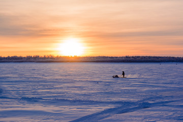 orange sky at sunset and blue snow with ice on the frozen river on which a man rides on a sleigh