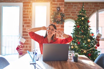 Beautiful woman sitting at the table working with laptop at home around christmas tree Doing peace symbol with fingers over face, smiling cheerful showing victory