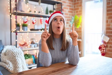 Young beautiful woman wearing christmas hat sitting at the table at home amazed and surprised looking up and pointing with fingers and raised arms.