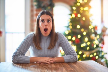 Obraz premium Young beautiful woman sitting at the table at home around christmas tree and decoration afraid and shocked with surprise expression, fear and excited face.