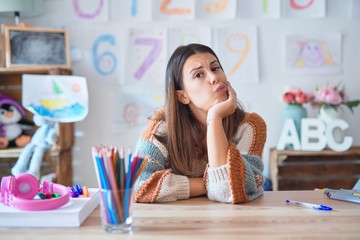 Young beautiful teacher woman wearing sweater and glasses sitting on desk at kindergarten thinking looking tired and bored with depression problems with crossed arms.