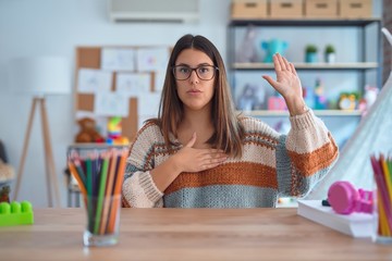 Young beautiful teacher woman wearing sweater and glasses sitting on desk at kindergarten Swearing...