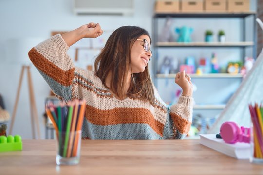 Young beautiful teacher woman wearing sweater and glasses sitting on desk at kindergarten Dancing happy and cheerful, smiling moving casual and confident listening to music