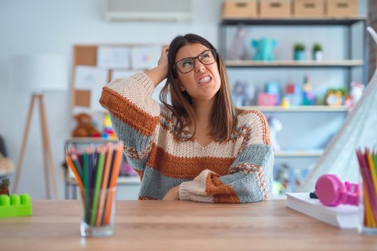 Young beautiful teacher woman wearing sweater and glasses sitting on desk at kindergarten confuse and wondering about question. Uncertain with doubt, thinking with hand on head. Pensive concept.