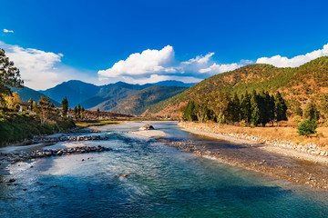 Naklejka na ściany i meble Mo Chhu River on a nice sunny day, Punakha, Bhutan. View from the wooden cantilever bridge near Punakha Dzong to river, houses of Punakha city and Himalaya mountains covered with forest.