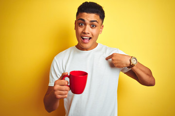 Young brazilian man drinking cup of coffee standing over isolated yellow background with surprise face pointing finger to himself