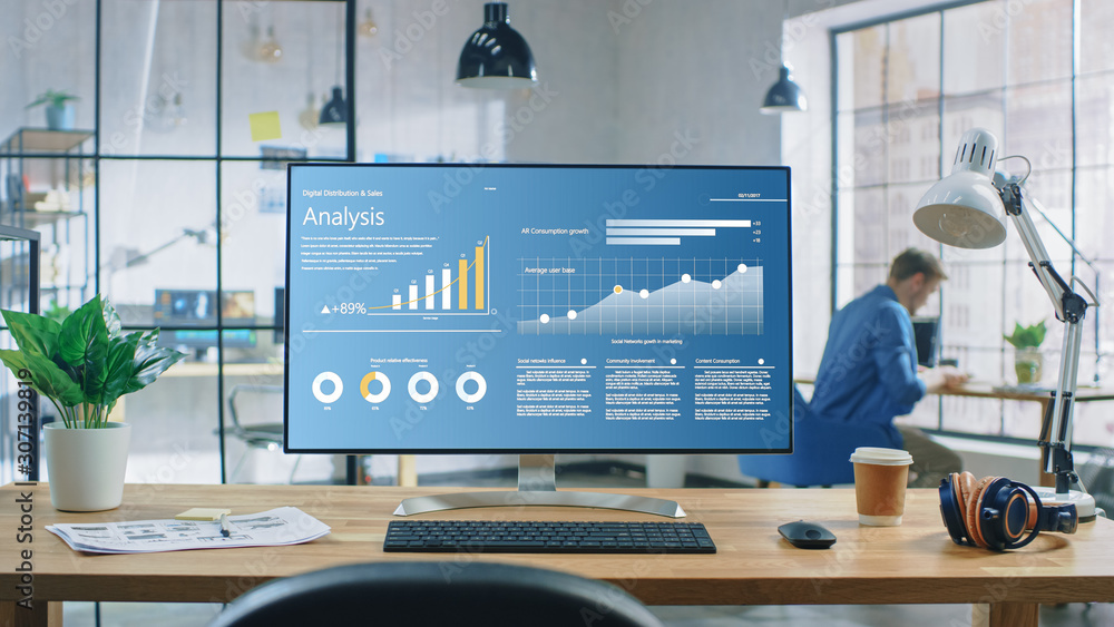 Poster Shot of a Desktop Computer in the Creative Modern Office. Monitor Screen Shows Company Growth Data with Graphs, Charts, Software UI. In the Background Young Professional Using Smartphone.