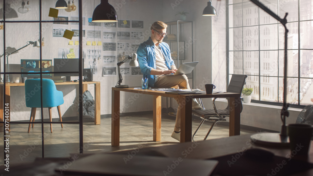 Poster professional creative designer sits on his desk holds laptop on the knees early morning while workin