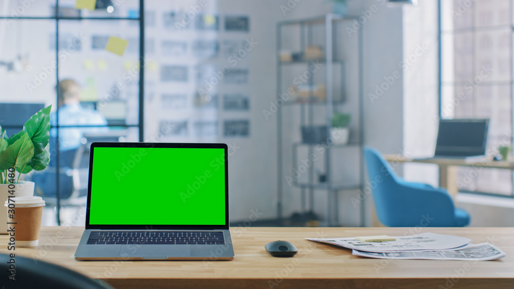 Poster laptop on the desk in the office shows green mock-up screen. in the background creative office or bu