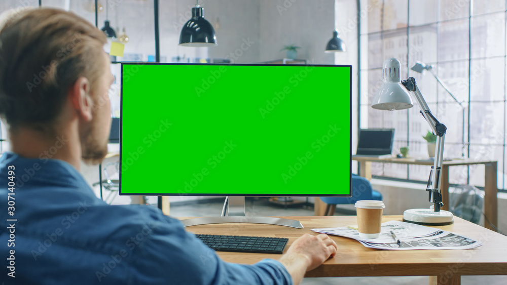 Aufkleber Over the Shoulder Shot: Man Uses Desktop Computer with Green Mock-up Screen while Sitting at His Desk. In the Background Stylish Modern Open Space Creative Business Office